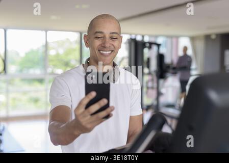 Bald Hispanic man working out on a stationary exercise bike inside modern gym while using phone. Focused fitness and workout concept showing strength Stock Photo