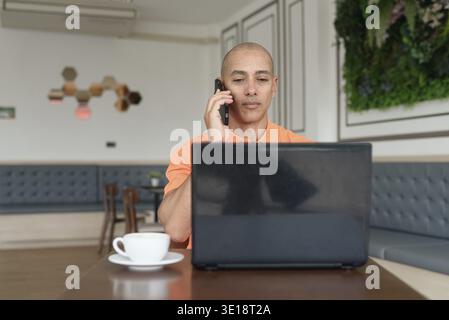 Handsome middle aged bald Hispanic man in orange polo shirt at cafe table with coffee cup working on laptop computer and talking on phone indoors. Rem Stock Photo