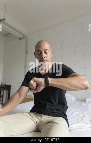 Middle aged bald Hispanic man in black t-shirt sitting on bed while looking at smartwatch. Indoor lifestyle concept in minimalistic apartment with mod Stock Photo