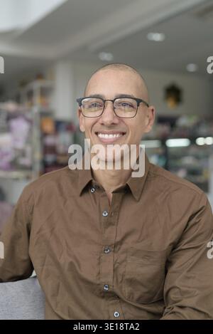 Bald Hispanic man in his 40s sitting in coffee shop, wearing casual shirt and eyeglasses. Confident and happy expression. Urban lifestyle, modern man Stock Photo