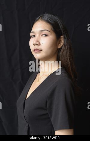 Young Asian woman in black dress posing against black curtain in natural light Stock Photo