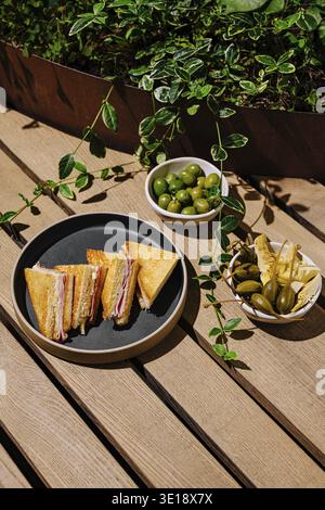 A platter of freshly cut sandwiches sits on a wooden table surrounded by greenery. Small bowls of olives and pickles are placed alongside, adding to t Stock Photo