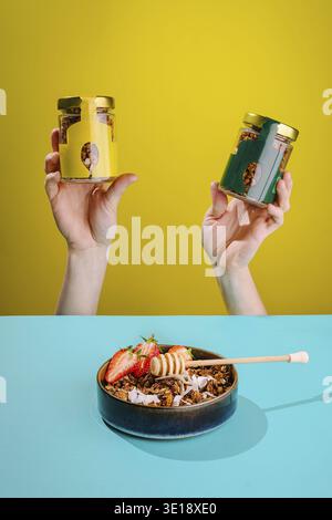 Two hands hold jars of nutritious snacks above a bowl filled with granola, fresh strawberries, and honey on a vibrant table setting during the day Stock Photo