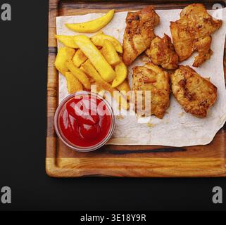 Crispy fried chicken pieces are served alongside golden fries and a small bowl of ketchup, arranged neatly on a wooden tray with parchment paper Stock Photo