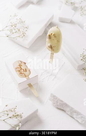 Two ice cream bars on sticks are placed on a smooth white surface. Surrounding elements include small white stones and delicate flowers, creating a cl Stock Photo