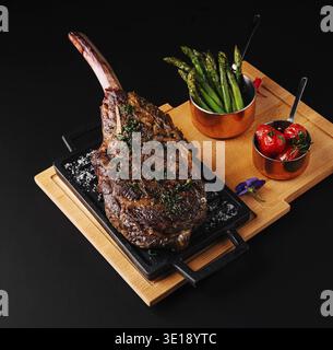 A large tomahawk steak cooked to perfection rests on a sizzling platter, garnished with herbs. Accompanying it are grilled asparagus and cherry tomato Stock Photo