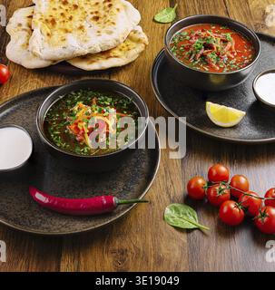 Two bowls of flavorful lentil soups topped with fresh herbs sit on a wooden table beside naan bread and fresh tomatoes, creating a warm and inviting d Stock Photo