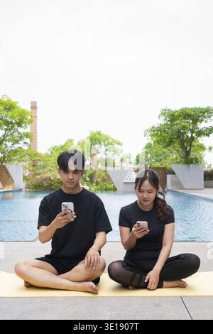 Young sporty couple doing yoga and using phone together next to swimming pool Stock Photo
