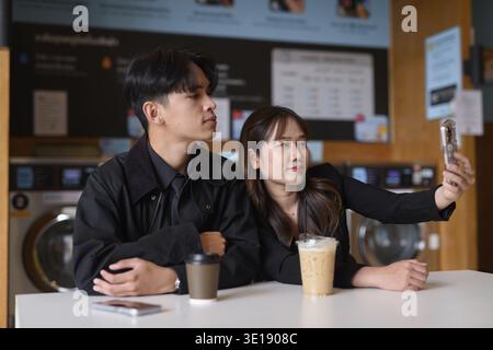 Young happy Asian couple sitting in laundry shop drinking coffee and using phone Stock Photo
