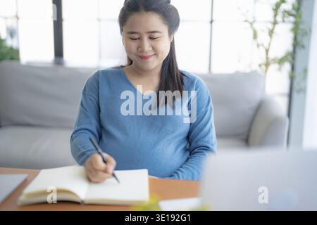 Pregnant asian woman writing notes and using a laptop while working at home pregnant businesswoman working on remote project in home office pregnancy Stock Photo