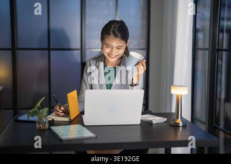 Alone Working Late at Night. Asian business woman at workplace in office late night work at a desk in a dark office working on a laptop late in the ev Stock Photo