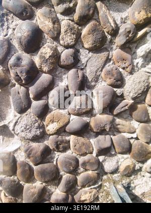 A vertical close-up shot of a textured stone wall featuring smooth, dark rounded rocks embedded in light-colored mortar. The natural afternoon light h Stock Photo
