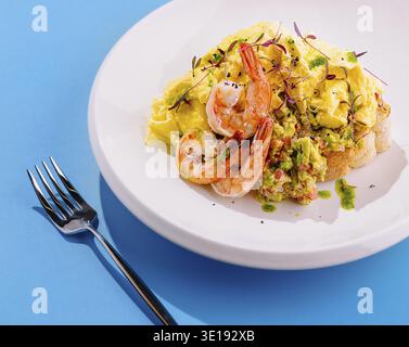 Scrambled eggs with shrimp and avocado on toast seasoned with microgreens and spices served on white plate over blue background Stock Photo