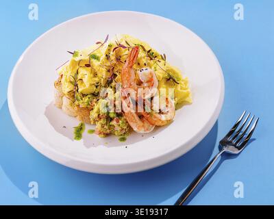 Scrambled eggs with shrimps served on toasted bread with mashed avocado and herbs on white plate over blue background Stock Photo