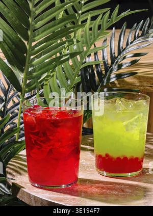 Two colorful cocktails with ice cubes are standing on the bar counter with tropical plants on the background Stock Photo