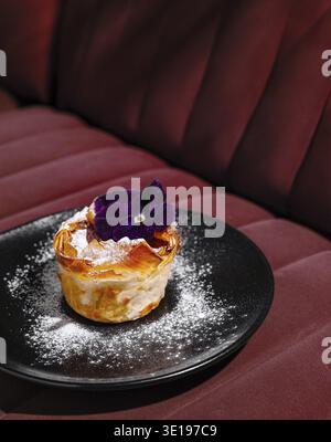 Delicious apple tart with a purple edible flower garnish, presented on a sophisticated black plate against a red backdrop Stock Photo