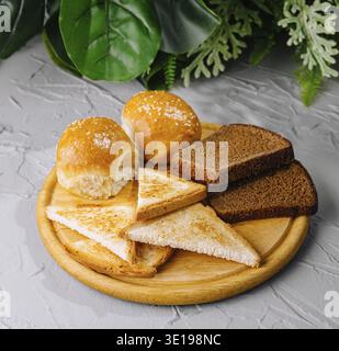 Variety of freshly baked breads on a round wooden cutting board, with green plants in background Stock Photo