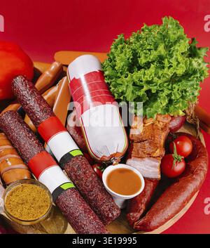 Appetizing composition of different types of sausages with herbs, spices and sauces on a wooden board, isolated on a red background Stock Photo