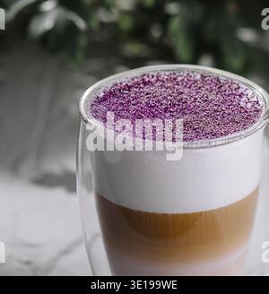 Trendy layered latte with purple foam in a clear mug, on a textured grey surface with greenery Stock Photo