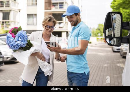 Young and professional delivery man in blue uniform delivers flowers to a woman client Stock Photo