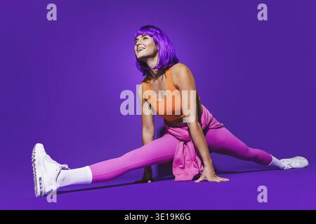 Young cheerful woman wearing colorful sportswear doing splits against purple background Stock Photo
