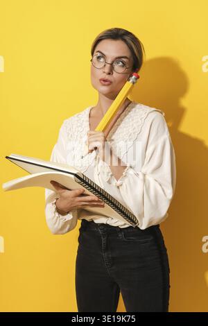 Thoughtful young woman wearing glasses holding big pencil and notebook on yellow background Stock Photo