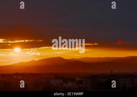 The sun sets behind hazy mountains, casting a warm orange glow over a silhouetted city skyline and residential buildings. Stock Photo