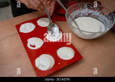 Person's hands carefully filling a red silicone mold with white mochi dough using a spoon, preparing traditional japanese confectionery in a home kitchen, showcasing the cooking process Stock Photo
