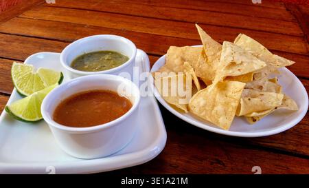 Traditional Mexican tortilla chips served with red and green chili sauces and fresh lime wedges on a natural wooden table. Stock Photo