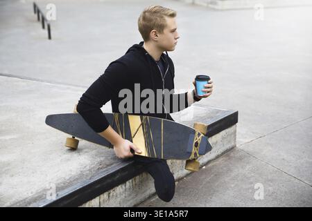 Young and motivated handicapped guy with a cup of coffee before longboard riding in a skatepark Stock Photo