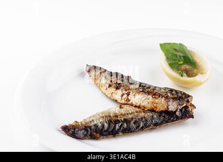 Two grilled mackerel fillets are served on a white plate with a lemon and basil garnish Stock Photo