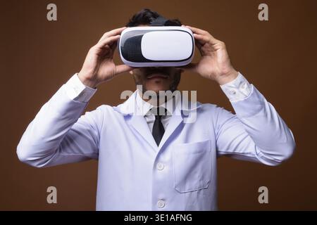 Studio shot of young handsome Hispanic man doctor using virtual reality headset against brown background Stock Photo