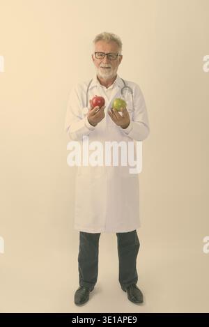 Full body shot of happy senior bearded man doctor smiling while holding red apple and green apple against white background Stock Photo