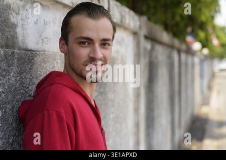 Young happy bearded man smiling while wearing red hoodie and leaning on old cement wall in the streets outdoors Stock Photo