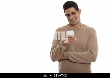 Studio shot of young happy Persian man smiling while using mobile phone Stock Photo