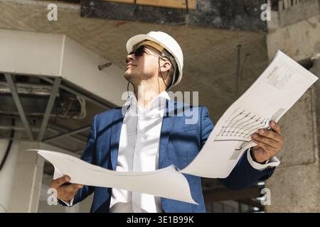 Young man architect wearing formal suit and hard hat during building construction control holding a blueprints on a construction site Stock Photo