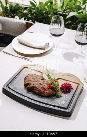 A beautifully presented grilled steak is placed on a hot stone, accompanied by garnishes. The dining table is elegantly set with wine glasses and gree Stock Photo