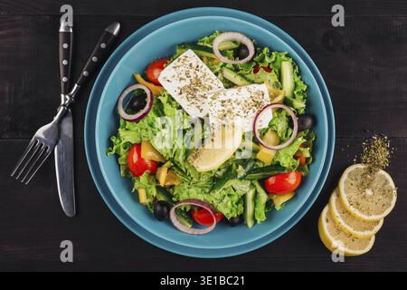 Greek salad with feta cheese, tomatoes, cucumbers, olives, oregano, and lemon juice in a blue bowl on a dark wooden table Stock Photo