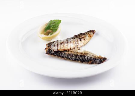 Two grilled mackerel fillets are served on a white plate with a lemon and basil garnish Stock Photo