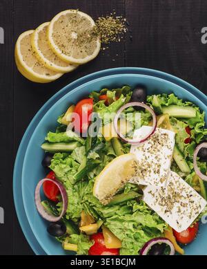 Greek salad with feta cheese, tomatoes, cucumbers, olives, oregano, and lemon juice in a blue bowl on a dark wooden table Stock Photo