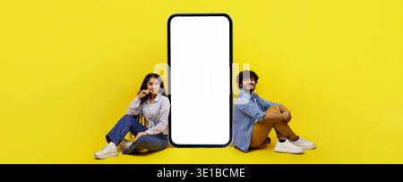 Couple Sits on the Floor Next to a Blank Smartphone Display in a Bright Yellow Setting Stock Photo