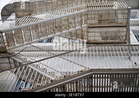 A professional shot featuring a complex, multi-level metal fire escape staircase attached to a white brick wall. The perspective highlights the rhythm Stock Photo