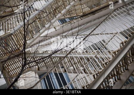 A professional shot featuring a complex, multi-level metal fire escape staircase attached to a white brick wall. The perspective highlights the rhythm Stock Photo