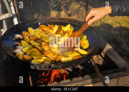 Frying potatoes on outdoor fire griddle. Close-up of sliced potatoes frying on a large outdoor griddle over open fire, showing rustic cooking, camping Stock Photo