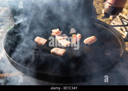 Pork fat frying on smoky outdoor griddle. Close-up of sliced pork fat cooking on a hot metal griddle with rising smoke, showing rustic outdoor food Stock Photo