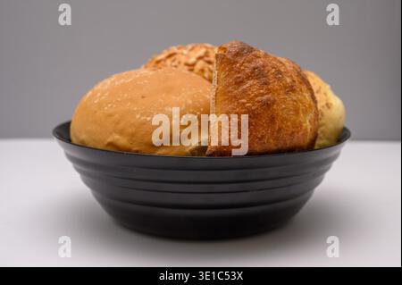 Various types of bread rolls rest in a black bowl on a clean surface. The rolls show different shapes and textures, creating a simple display of baked Stock Photo