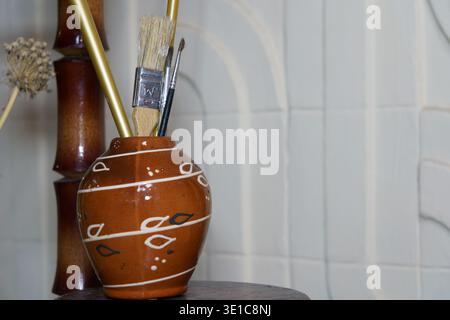 Brushes and tools in a brown vase on a wooden table in an art studio setting Stock Photo