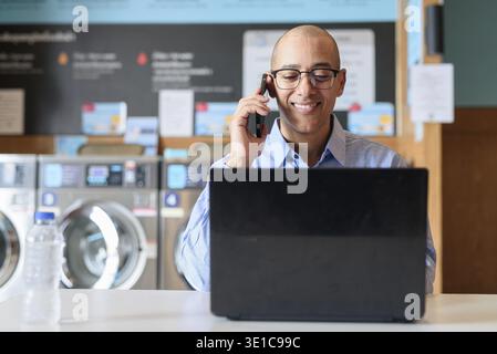 Bald male professional in blue button-down shirt working on laptop inside laundry shop. He is using technology in everyday environment. Real life work Stock Photo