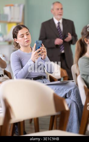 Young female student scans QR code or takes photo, teacher in background Stock Photo