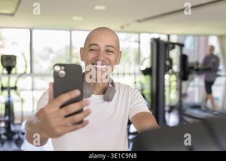 Bald Hispanic man working out on a stationary exercise bike inside modern gym while using phone. Focused fitness and workout concept showing strength Stock Photo